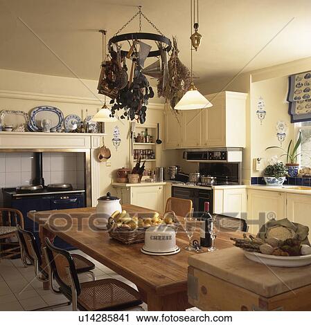 Pendant light above chrome and wicker chairs and rectangular wooden table in cream country kitchen with utensils on circular metal rack View Large Photo Image Stock Image - Pendant light above chrome and wicker chairs and rectangular wooden table in cream country kitchen with utensils on circular metal rack . Fotosearch