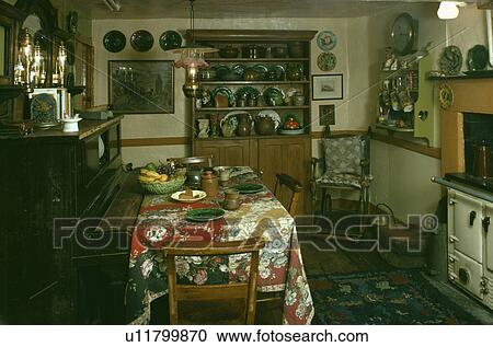 Stock Image - Piano in cottage kitchen with pine dresser and floral cloth on table in front of Aga oven. Fotosearch
