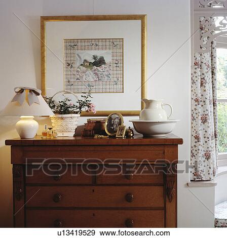 Stock Photo - Picture on wall above lighted lamp and jug and bowl on antique mahogany chest of drawers. Fotosearch