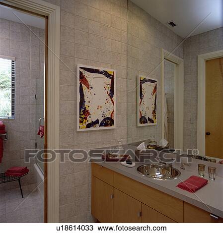 Stock Image - Picture on wall beside steel basin in grey and wood vanity unit below mirrored wall in beige tiled modern bathroom. Fotosearch