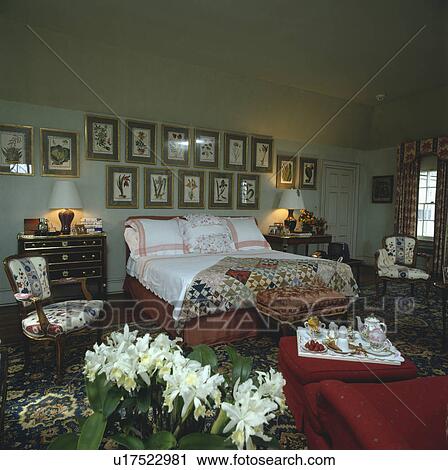 Stock Image - Pictures above bed with large white pillows and patchwork quilt in grey bedroom with breakfast tray on red chaiselongue. Fotosearch