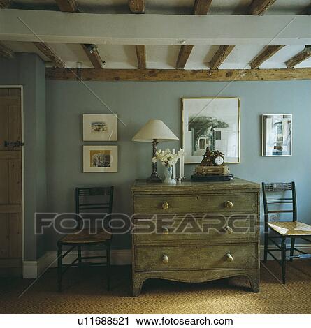 Stock Image - Pictures on blue wall above antique chest of drawers with white lamp. Fotosearch