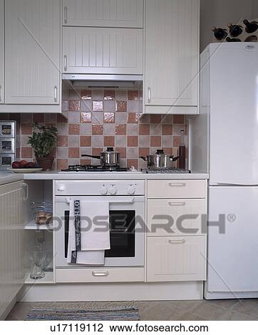 Stock Image - Pink+white tiles above white oven in small white kitchen with tall fridge-freezer. Fotosearch