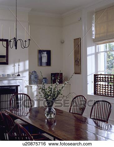 Polished antique table and Windsor chairs in white dining room with metal chandelier and cream blinds at window View Large Photo Image Stock Photograph - Polished antique table and Windsor chairs in white dining room with metal chandelier and cream blinds at window. Fotosearch