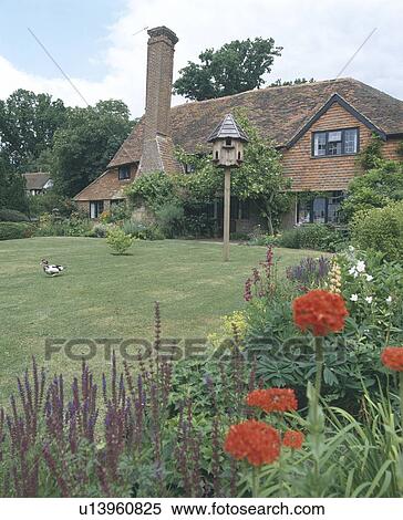 Stock Photography - Purple salvia and red lychnis in summer border beside lawn with dovecote in front of old red brick country house with tall chimney. Fotosearch