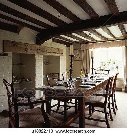 Stock Image - Rectangular wooden table and antique chairs in country dining room with beamed ceiling. Fotosearch