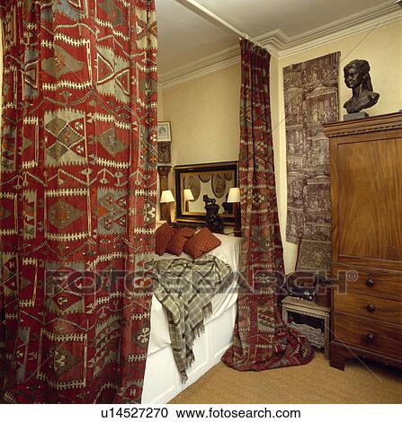 Stock Image - Red patterned kelim rugs on curtain rail above bed in traditional bedroom. Fotosearch