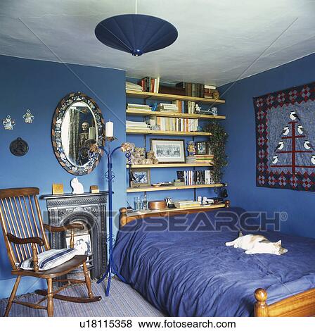 Rocking chair beside fireplace in blue bedroom with wooden shelves on wall behind bed with blue duvet View Large Photo Image Stock Photo - Rocking chair beside fireplace in blue bedroom with wooden shelves on wall behind bed with blue duvet. Fotosearch