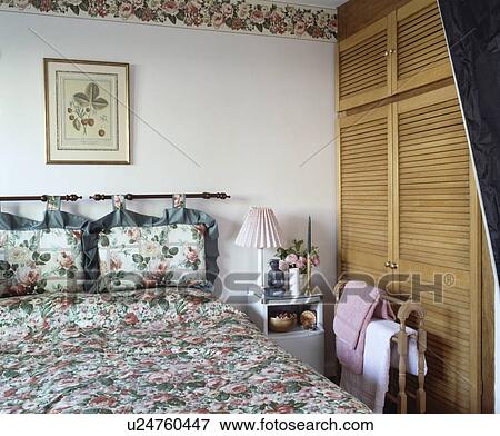 Rose-patterned cushions on pole above bed with matching quilt in small bedroom with wooden louvre doors on fitted unit View Large Photo Image Stock Photo - Rose-patterned cushions on pole above bed with matching quilt in small bedroom with wooden louvre doors on fitted unit. Fotosearch
