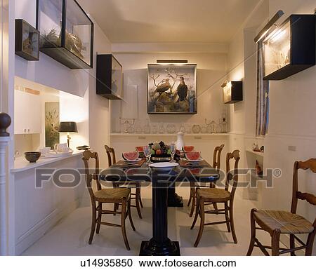 Stock Image - Rush-seated chairs and black pedestal table in narrow white dining room with stuffed birds in glass cases on wall. Fotosearch