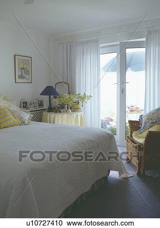 Stock Image - Seaside bedroom with white quilt on bed and white curtains at patio doors. Fotosearch