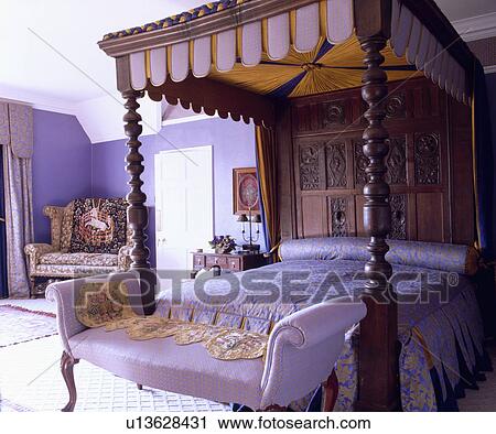 Stock Image - Small chaiselongue below antique Tudor carved wood fourposter bed in purple country bedroom. Fotosearch