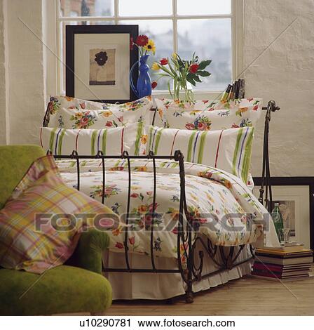 Stock Image - Striped and floral patterned pillows with floral quilt on wrought iron bed in front of window with picture on windowsill. Fotosearch