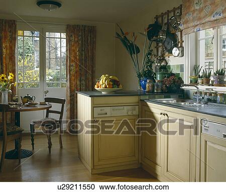 Stock Image - Traditional circular wood dining table and chairs in front of French windows in small openplan kitchen diningroom. Fotosearch