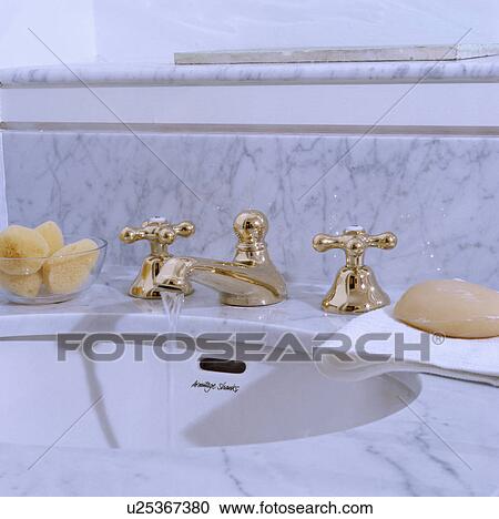 Stock Image - Water pouring from gold tap in white basin underset in marble unit. Fotosearch