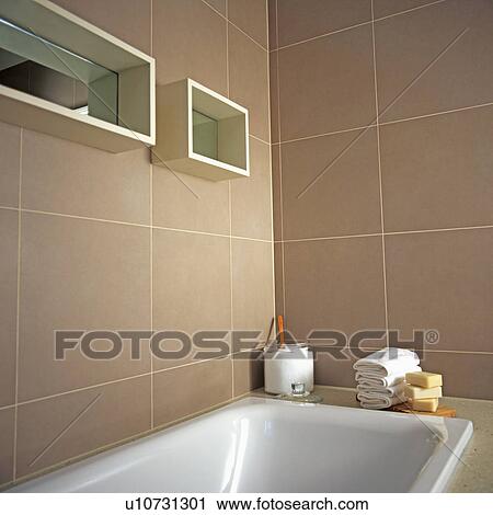 Stock Image - White bath below mirrored cube shelves on beige tiled walls of modern bathroom. Fotosearch