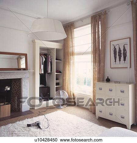 Stock Image - White chest-of-drawers and beige sheer curtains in modern white bedroom with hairdryer and electric fan on white rug&#13; &#10; &#13; &#10;. Fotosearch