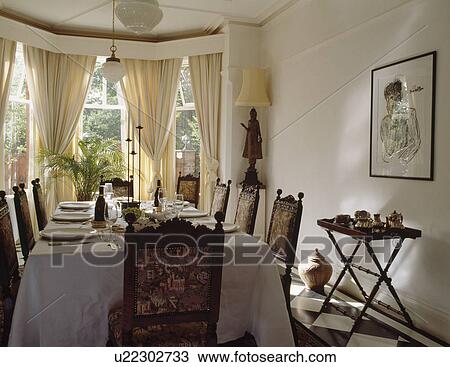 Stock Image - White cloth on table with upholstered chairs in white dining room with cream curtains. Fotosearch