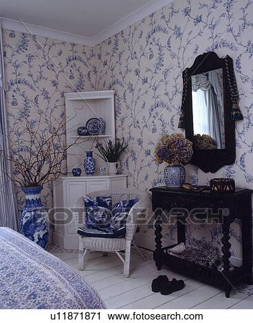 Stock Image - White corner cupboard and cane chair in bedroom with blue+white floral wallpaper and mirror above mahogany table. Fotosearch
