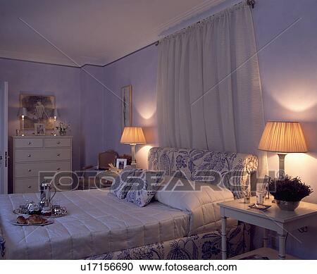 White drapes above blue+white upholstered bed with white bolster pillow in mauve bedroom with lighted lamps View Large Photo Image Stock Image - White drapes above blue+white upholstered bed with white bolster pillow in mauve bedroom with lighted lamps. Fotosearch
