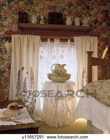 Stock Image - White embroidered curtains in cottage bedroom with jug on circular table with white cloth in front of window. Fotosearch