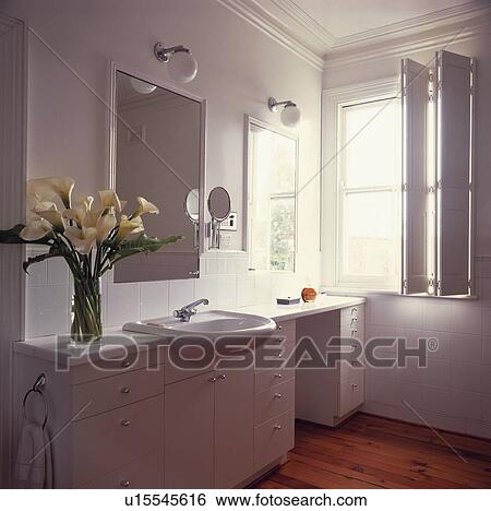 Stock Photograph - White lilies in vase on fitted vanity unit with basin in modern white bathroom with folding white shutters at window. Fotosearch