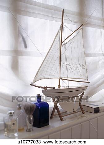 Stock Image - White model boat on window ledge against sheer white window covering . Fotosearch
