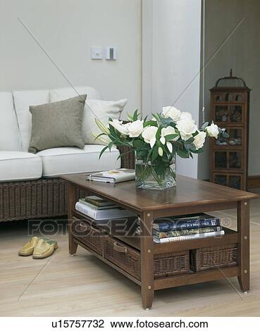 Stock Image - White paeonies on dark wood table in modern livingroom with white sofa and laminate wood floor. Fotosearch