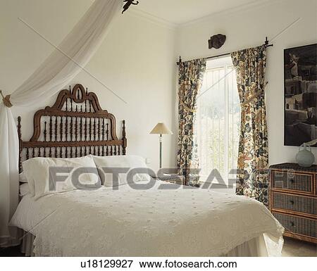 Stock Photo - White pillows and quilt on bed with carved wooded headboard in white bedroom with floral curtains. Fotosearch