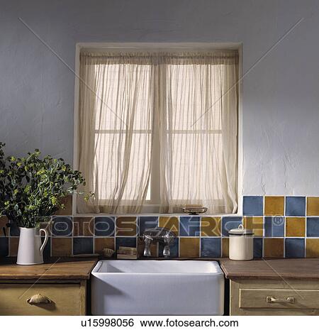 Stock Photograph - White sheer curtains and multicoloured tiles above white Butler's sink . Fotosearch