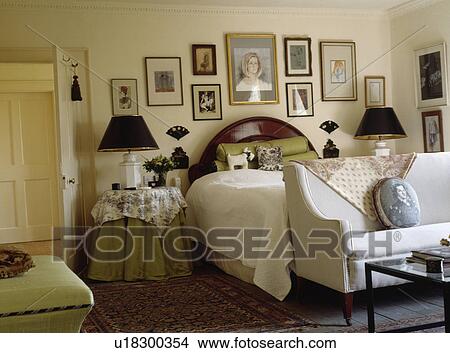 Picture - White sofa below bed in traditional bedroom with pictures on the walls and black lamps and cloths on bedside tables. Fotosearch