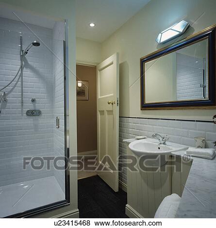 Stock Photo - White tiled shower with glass door in white bathroom with large mirror above basin. Fotosearch