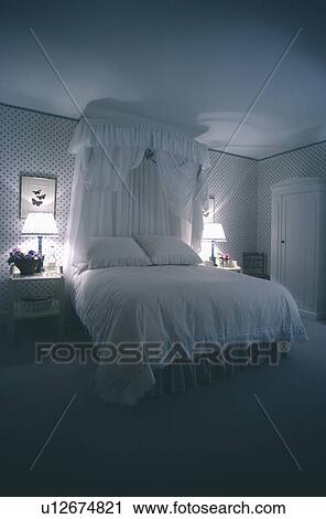 Stock Image - White voile drapes on half-tester bed with white linen in traditional bedroom with spotted wallpaper and lighted lamps. Fotosearch