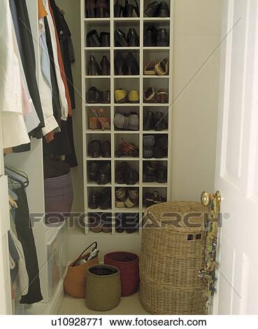 Stock Image - Wicker linen basket in front of shoes stored on cube shelves in walk-in wardrobe with hanging clothes. Fotosearch
