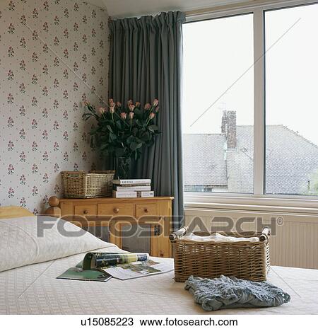 Stock Image - Wicker storage basket on bed in small bedroom with patterned wallpaper and green curtain. Fotosearch