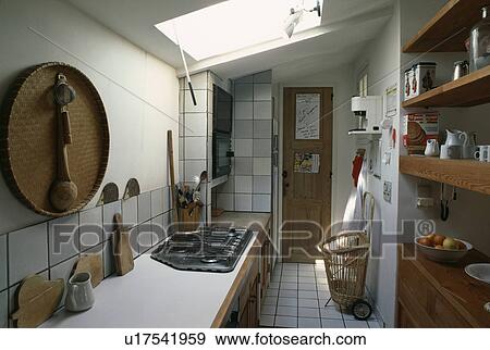 Stock Photo - Wicker tray on wall above worktop with built-in hob in white galley kitchen extension with open wooden shelves. Fotosearch