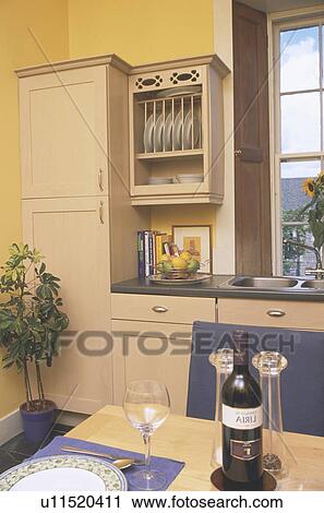 Stock Image - Wine bottle and settings on table in yellow kitchen with cream fitted cupboards. Fotosearch