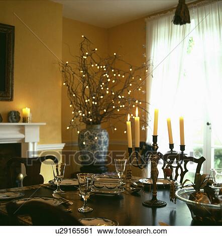 Stock Image - Yellow candles in candelabra on polished table in yellow dining room with lights on branches in pot in corner of room. Fotosearch