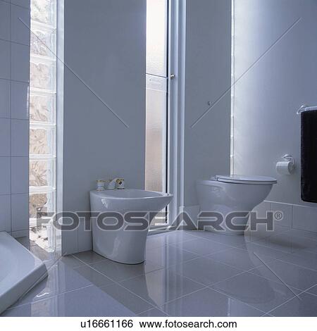 Stock Photograph - All white tiled architectural bathroom with bidet and louvred windows.. Fotosearch