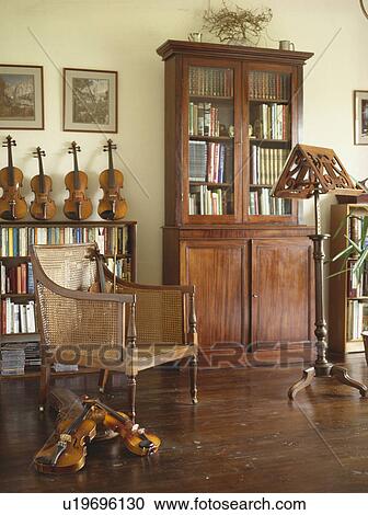 Stock Image - Antique bookcase and colonial cane chair in music room with wooden floor and collection of violins . Fotosearch