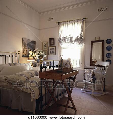 Stock Image - Antique Butler's Tray at the foot of brass bed in bedroom with white rocking chair and blue+white ribbon pattern quilt. Fotosearch
