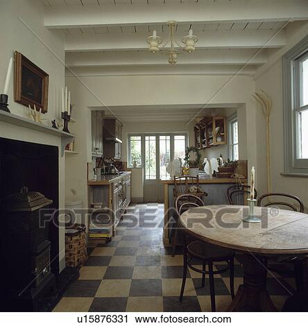 Stock Image - Antique pine table and Bentwood chairs in country kitchen dining room with chequerboard flooring and fireplace. Fotosearch