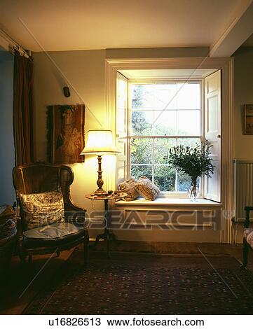 Stock Image - Armchair and lighted lamp beside window in traditonal cottage living room. Fotosearch