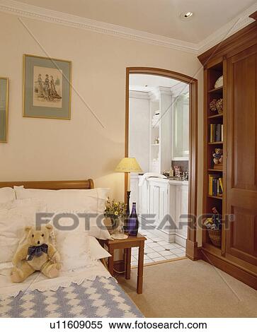 Stock Photography - Bed with white cushions and pillows and blue and white check quilt in bedroom with built-in cupboard and open door to ensuite bathroom. Fotosearch