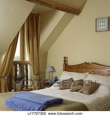 Stock Photography - Beige curtains at window in cream attic bedroom with beige cushions and cream linen on pine bed. Fotosearch