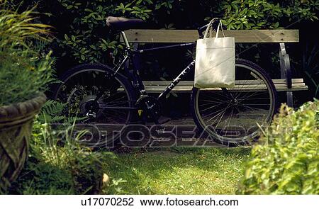 Stock Image - Bicycle with shopping bag leaning against old wooden bench in the garden. Fotosearch