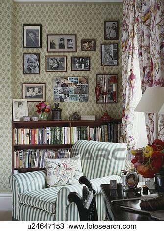Stock Image - Black and white photographs on wall above bookshelves in living room with green and white striped armchair. Fotosearch