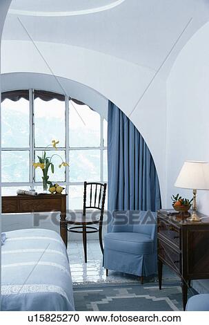Blue tubchair and antique cabinet beside arch with view of table and chair in front of window in white bedroom View Large Photo Image Stock Image - Blue tubchair and antique cabinet beside arch with view of table and chair in front of window in white bedroom. Fotosearch