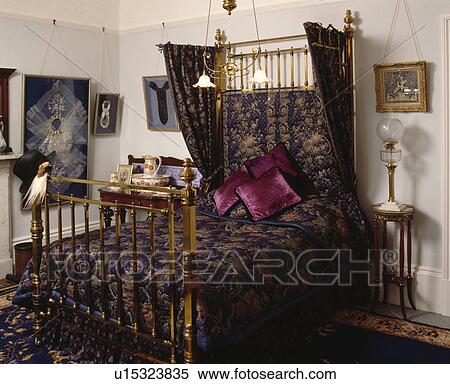 Stock Photography - Blue+gold drapes and quilt on Victorian brass half-tester bed in traditional bedroom. Fotosearch