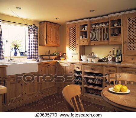 Stock Image - Blue+white checked curtains above white ceramic sink in cottage kitchen with fitted wooden cupboards. Fotosearch